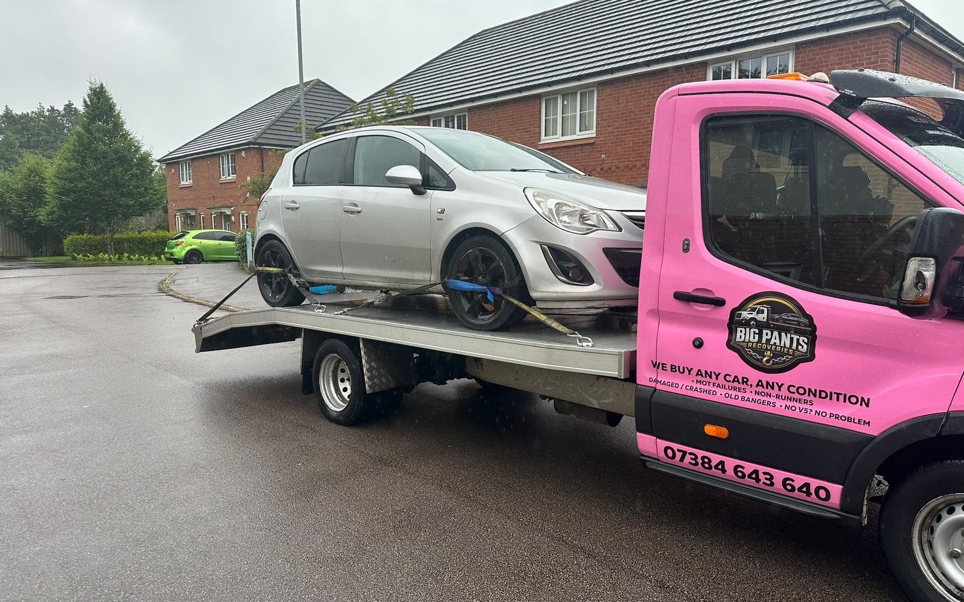A silver hatchback car strapped onto the flatbed of a pink 'Big Pants Recoveries' tow truck, parked on a residential street in front of brick houses.