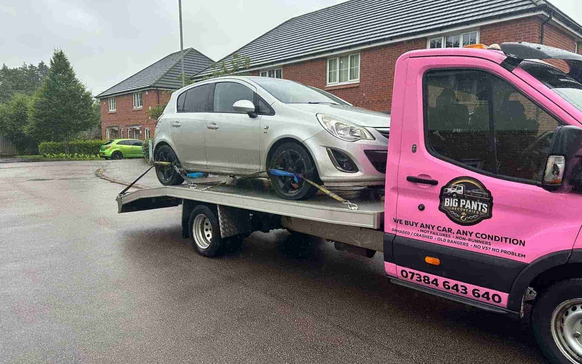 A silver hatchback car strapped onto the flatbed of a pink 'Big Pants Recoveries' tow truck, parked on a residential street in front of brick houses.